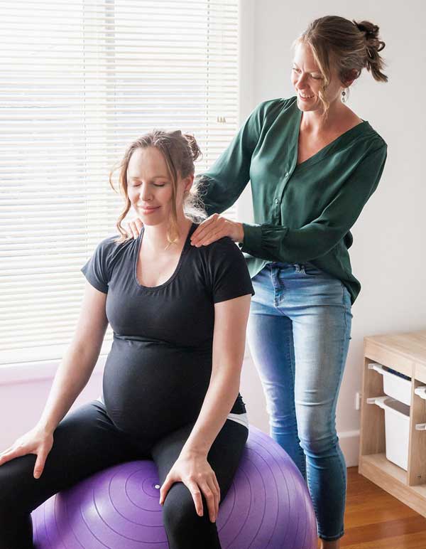 Doula standing behind pregnant mother who is sitting on an exercise ball giving her a shoulder massage